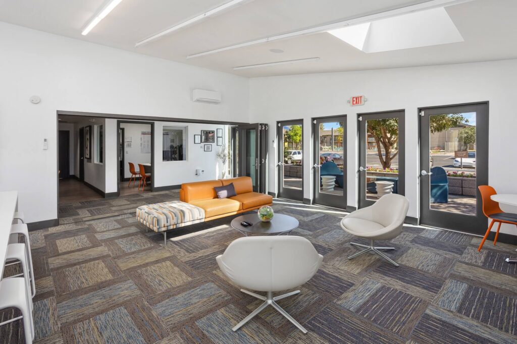 Modern lounge area with a brown leather sofa, round table, white chairs, and large windows letting in natural light. Gray patterned carpet and white walls give the space a clean, contemporary look.