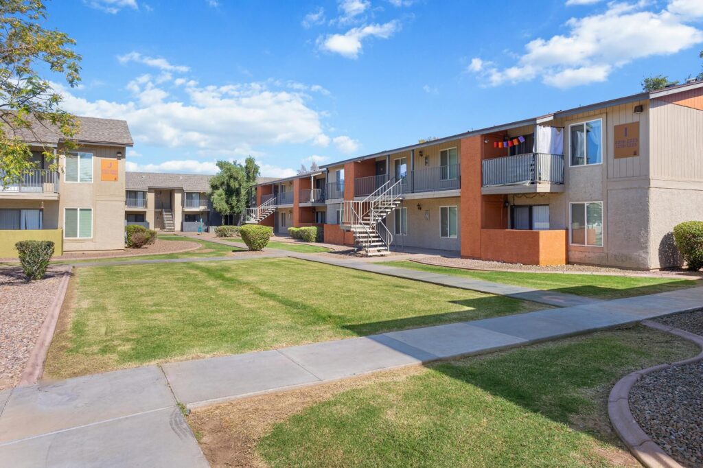Two-story apartment buildings with beige and orange accents, surrounded by trimmed bushes and a grassy courtyard, under a blue sky with scattered clouds. Concrete walkways lead through the landscaped area.