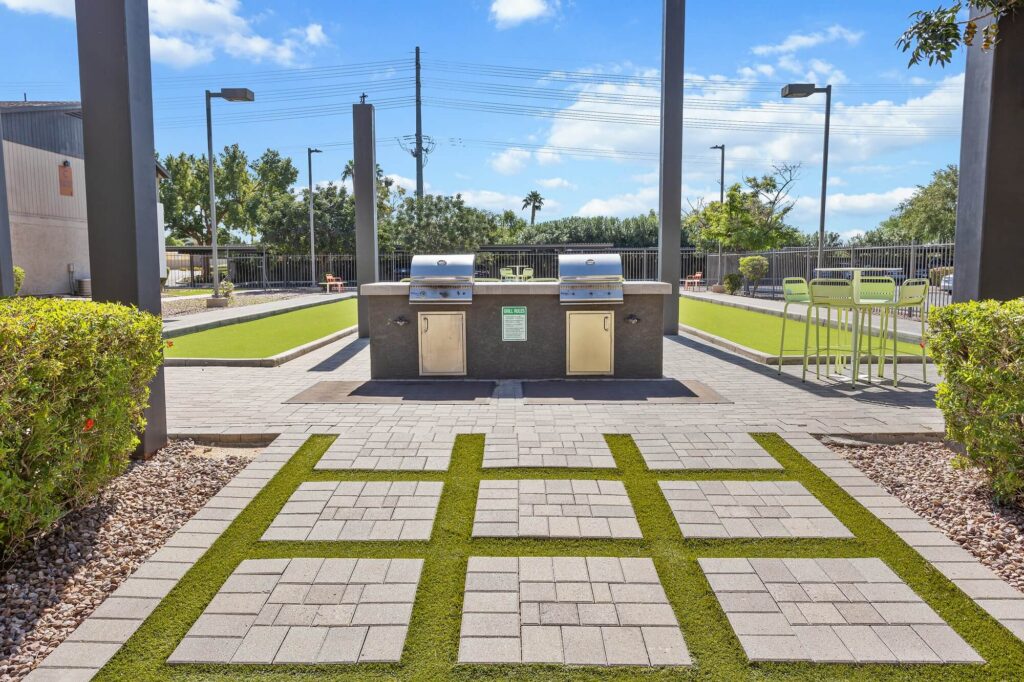 Modern outdoor patio with two stainless steel grills, green turf accents between pavers, bar-height tables and chairs, and manicured bushes under a blue sky with scattered clouds.