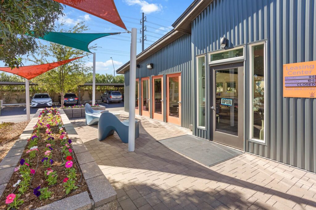Leasing center exterior with a glass door reading OPEN, colorful shade sails above, a flower bed, modern benches, and parked cars in the background on a sunny day.