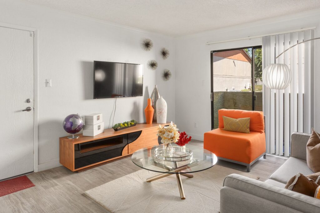 Modern living room with an orange chair, glass coffee table, TV on the wall, wooden TV stand, decorative vases, and wall art. Sliding glass doors let in natural light.