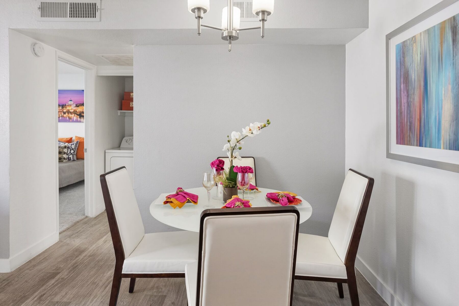 A round dining table with four white chairs is set with pink napkins, flowers, and glasses. A chandelier hangs above, and a colorful abstract painting decorates the wall. A bedroom is visible through an open doorway.