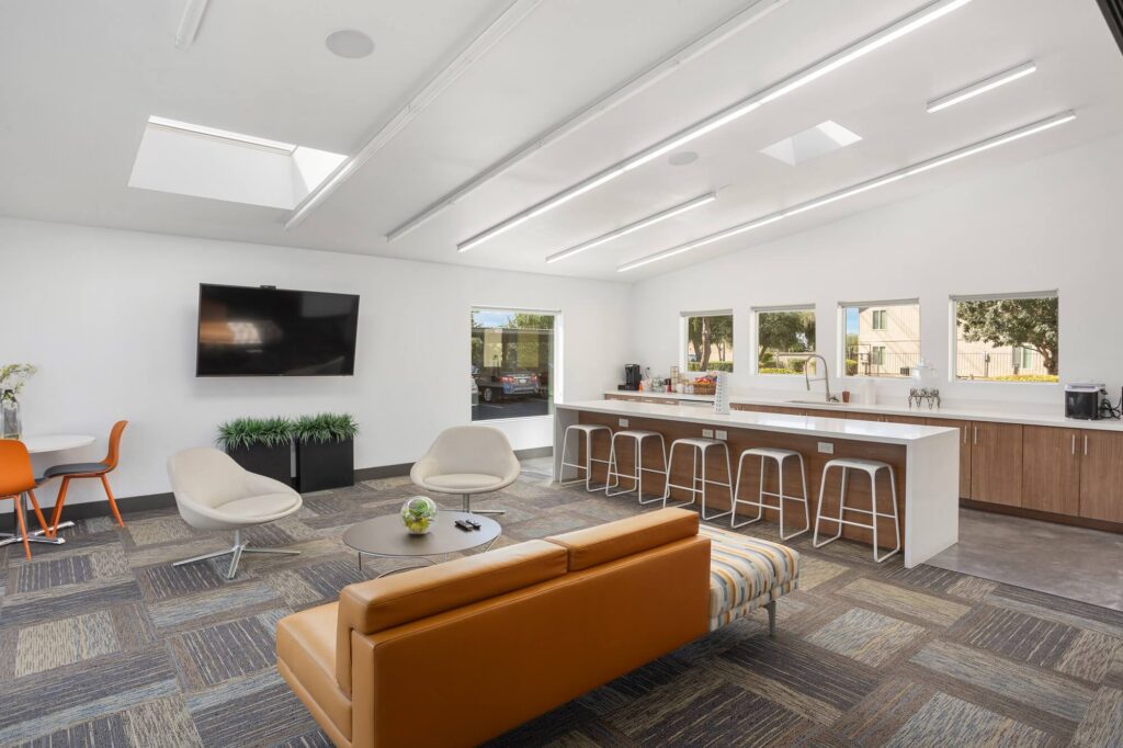Modern lounge with an orange sofa, chairs, wall-mounted TV, kitchenette with barstools, and windows. Neutral colors, patterned carpet, skylights, and a mix of natural and artificial lighting enhance the space.