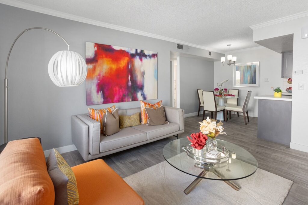 Modern living room with a gray sofa, orange accents, abstract wall art, glass coffee table with flowers, dining area with a white table and four chairs, and a contemporary floor lamp. The space is bright and stylish.