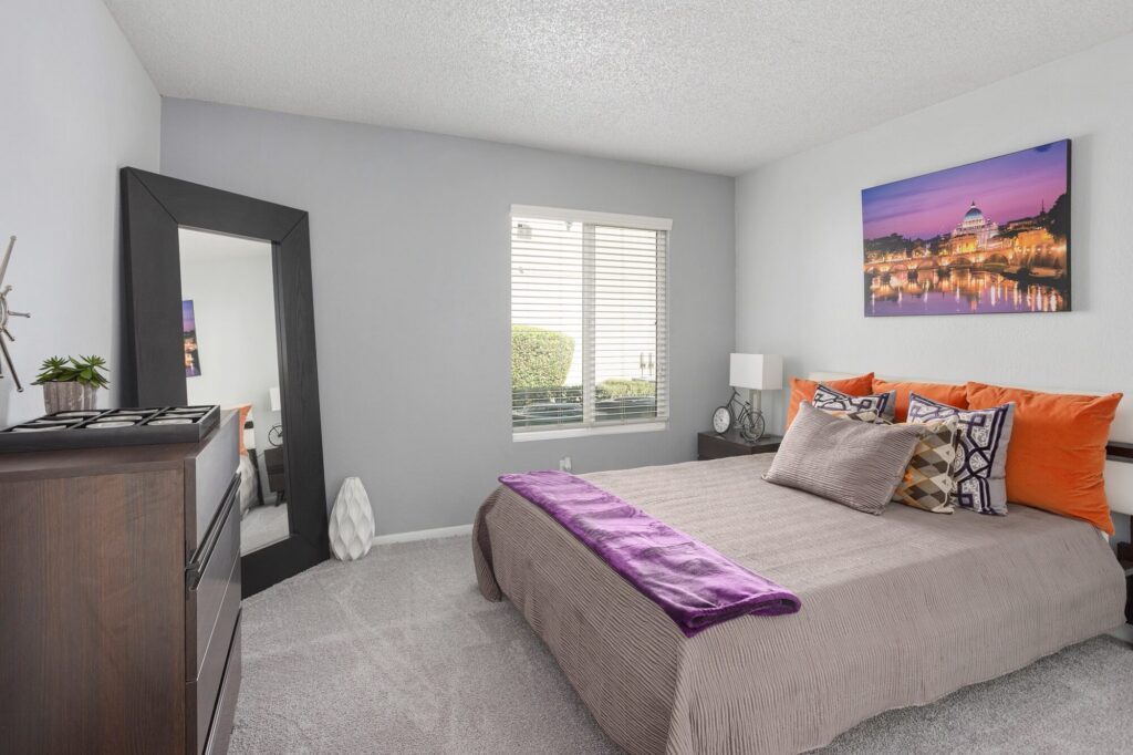A neatly made bed with orange and patterned pillows, a purple throw, a dresser, a tall mirror, a window with blinds, and a cityscape artwork on a gray wall in a bright, tidy bedroom.