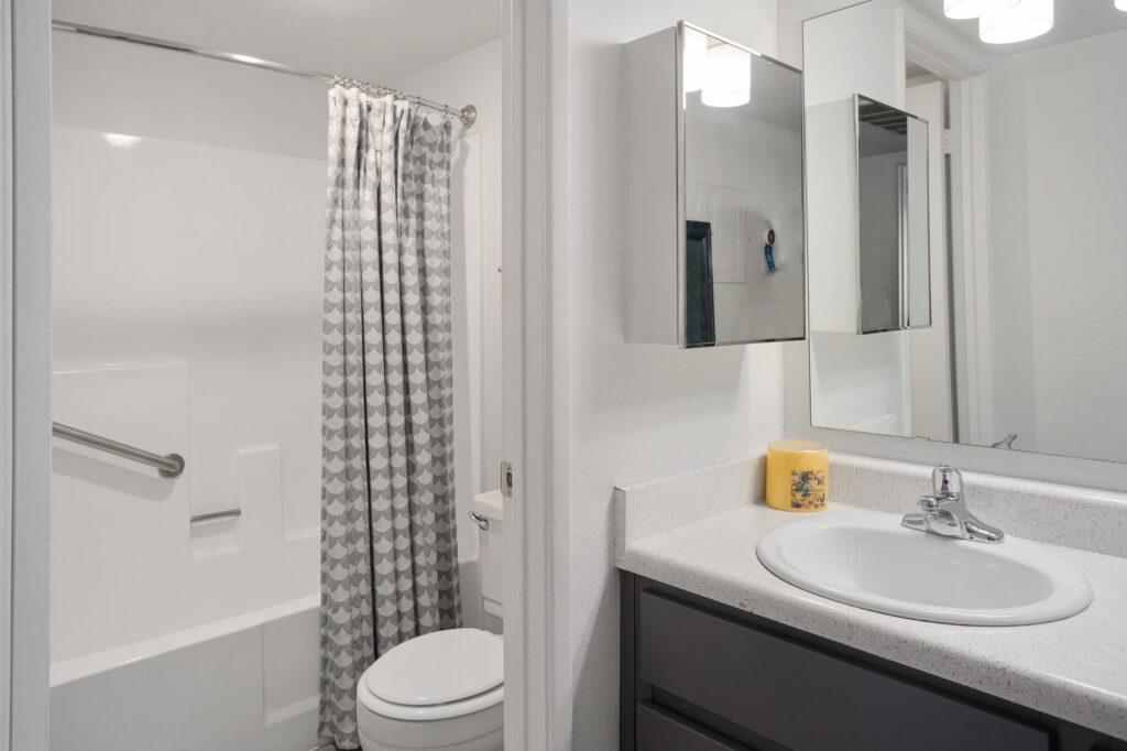 Modern bathroom with a white bathtub, gray-patterned shower curtain, toilet, sink with countertop, wall-mounted mirror cabinet, and a yellow candle. The space is clean and well-lit with white walls.