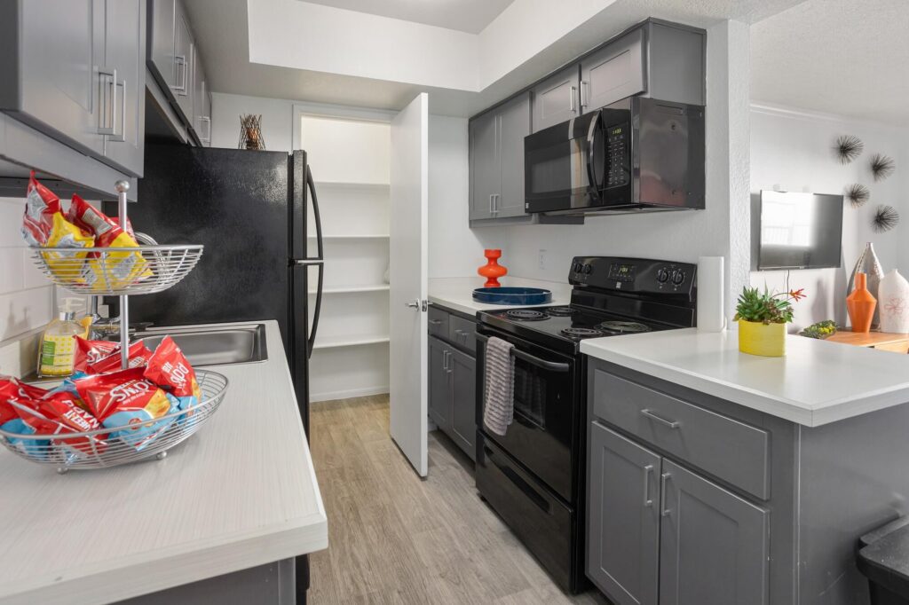 Modern kitchen with gray cabinets, black appliances, white countertops, and pantry door open. A tiered basket with snacks sits on the counter, with a small plant and orange vase for decor.