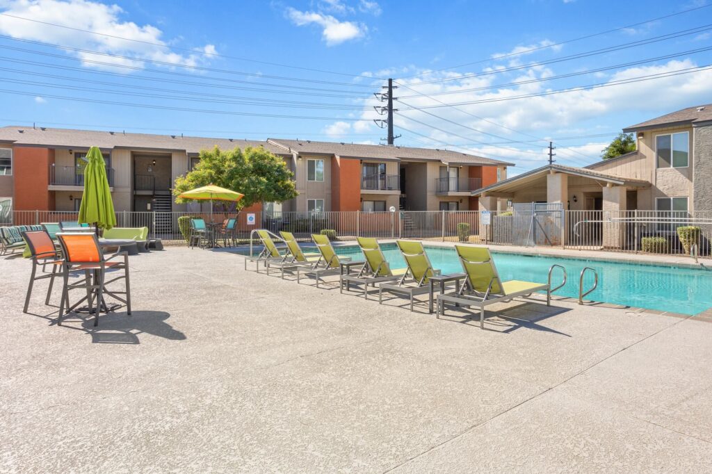 Outdoor apartment pool area with green lounge chairs, tables with umbrellas, and a fenced perimeter. Apartment buildings and power lines are visible in the background under a sunny blue sky.