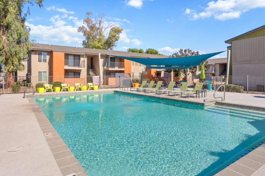 Outdoor apartment pool with clear blue water, surrounded by lounge chairs and green seating. A sunshade covers part of the deck. Two-story apartment buildings and trees are in the background under a blue sky.