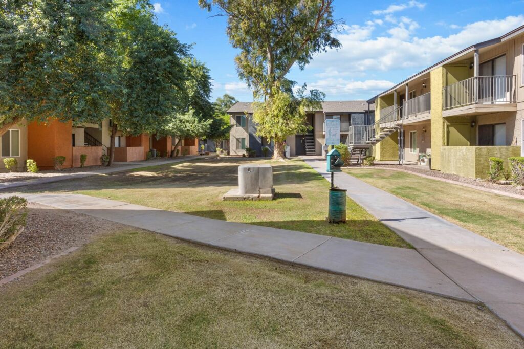 A courtyard view of an apartment complex with two-story buildings, grass, trees, a sidewalk, and a green trash can under a sunny sky.