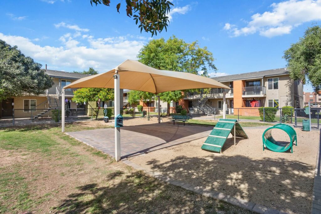 A shaded outdoor area in an apartment complex features a play zone with ramps and a tunnel for dogs, surrounded by grass, trees, and apartment buildings under a blue sky with clouds.