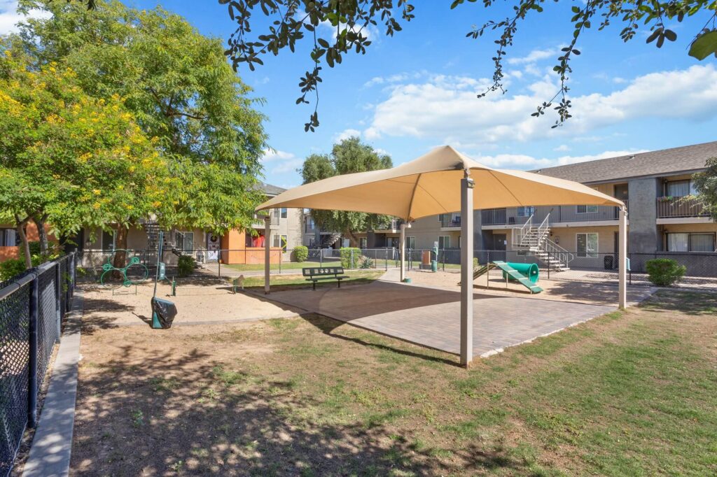 A playground with a large shade canopy, sand area, climbing structures, benches, and apartment buildings in the background, surrounded by grass, trees, and a chain-link fence.