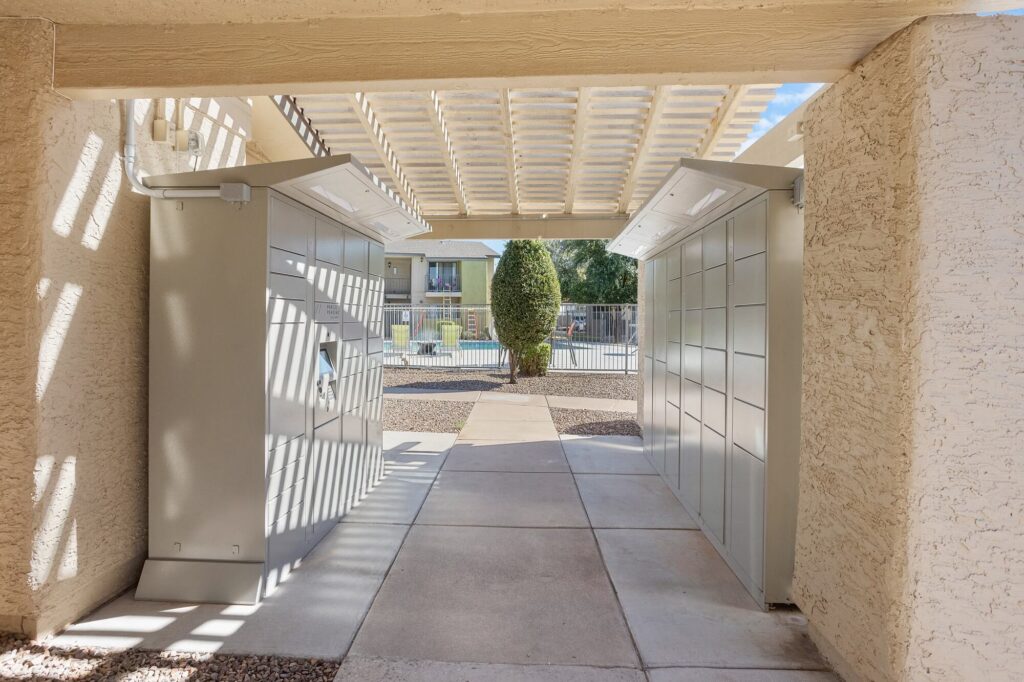 Two open metal package lockers stand under a covered walkway, casting shadows on the ground. In the background, there is a tree, fence, and buildings on a sunny day.