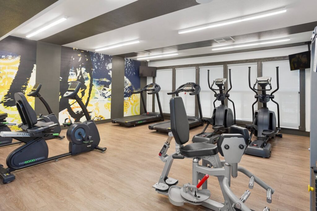 A modern gym with exercise bikes, treadmills, elliptical machines, and a weight machine on a wood floor. Large windows and a colorful mural decorate the space.