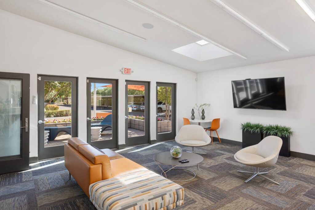 Modern lounge with tan couch, two white chairs, round table, wall-mounted TV, and large windows letting in natural light. Small desk and chair in corner, indoor plants, and view of outdoor seating area.