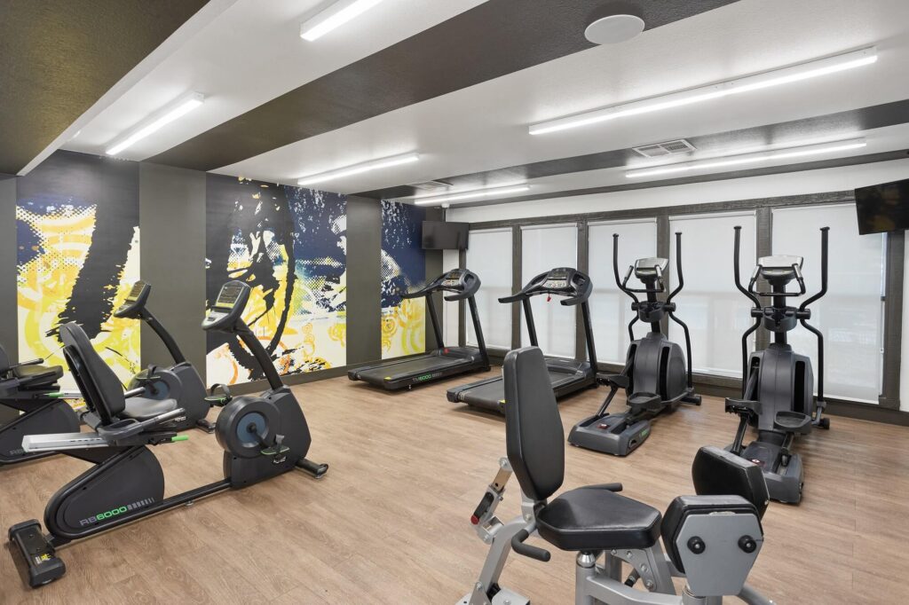 Modern gym room with wood floors, exercise bikes, elliptical machines, and treadmills. Large windows line one wall; vibrant abstract wall art decorates the space. Bright overhead lights illuminate the area.
