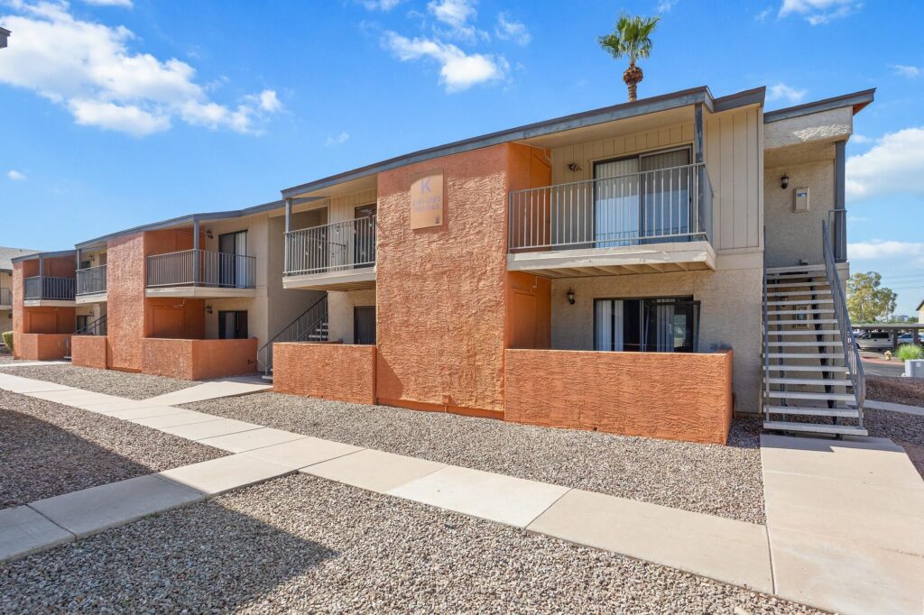 Two-story apartment building with orange and beige walls, balconies, and exterior staircases. The area is landscaped with gravel and has concrete walkways. A single palm tree is visible behind the roof. Blue sky with clouds.