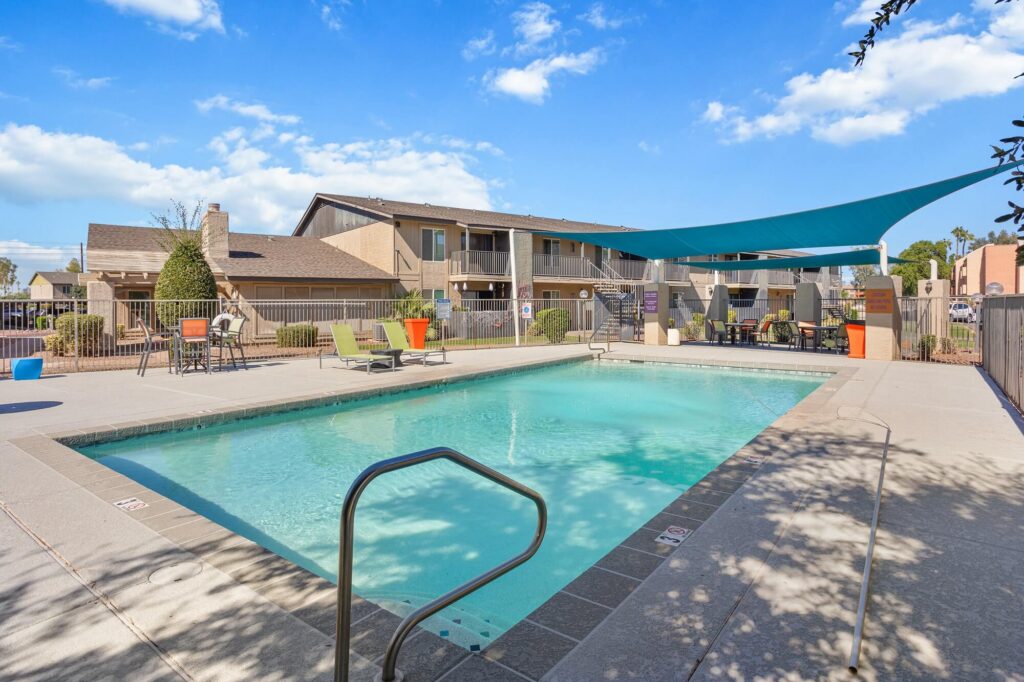 Outdoor swimming pool with clear water, surrounded by lounge chairs and tables, next to an apartment building under a blue sky with scattered clouds. A blue sunshade covers part of the pool deck.
