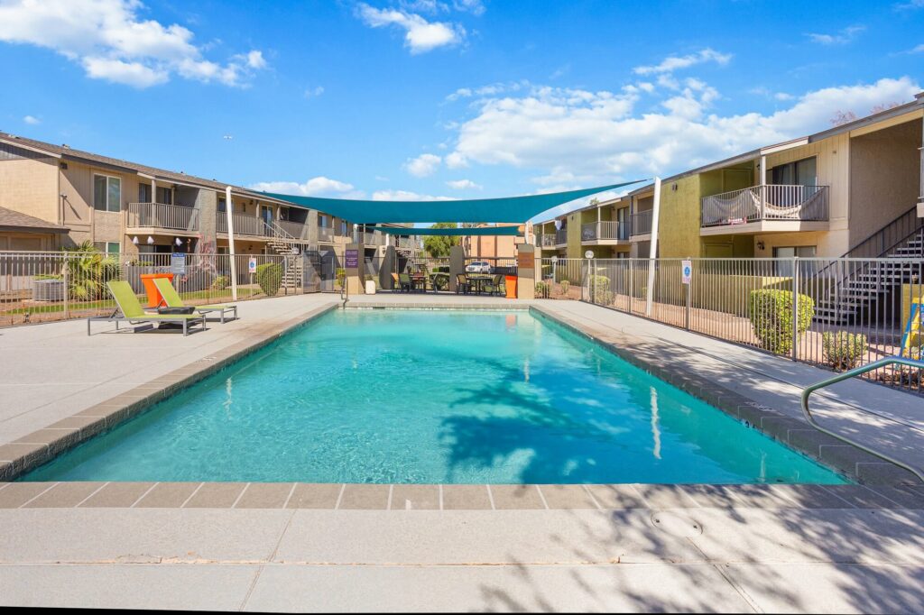 A rectangular outdoor swimming pool with lounge chairs and a blue shade canopy, surrounded by apartment buildings and a clear blue sky with scattered clouds.