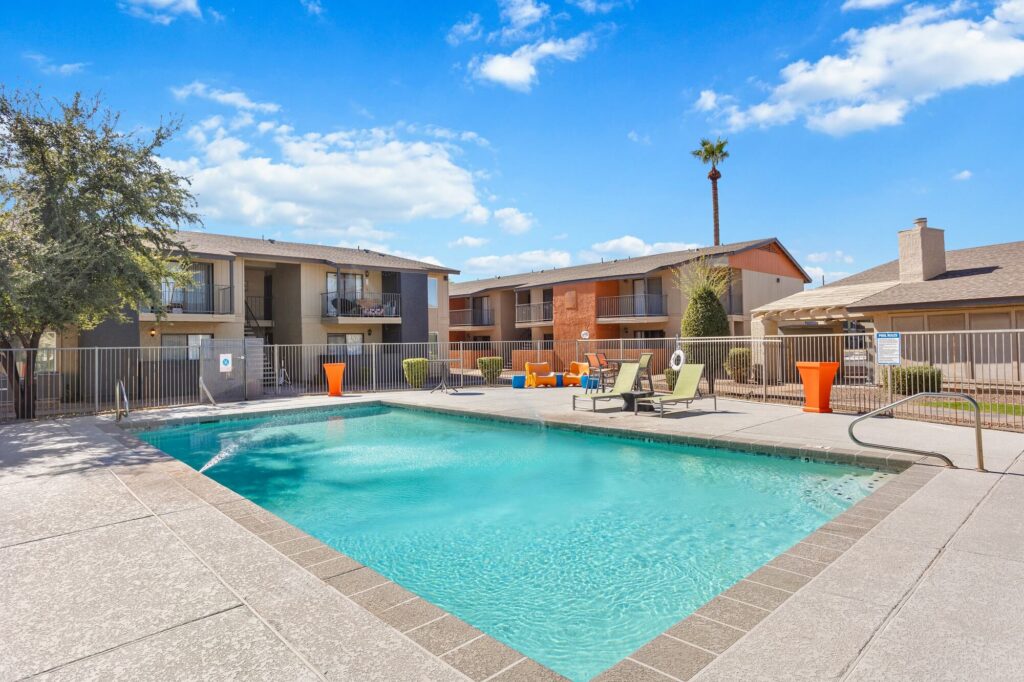 A rectangular outdoor swimming pool surrounded by lounge chairs, orange planters, and apartment buildings under a blue sky with scattered clouds.