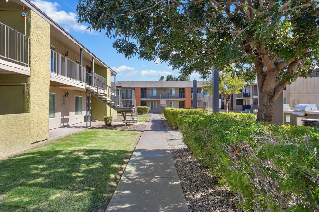 A sidewalk runs between two beige apartment buildings, bordered by green grass, bushes, and a large leafy tree under a blue sky with clouds.