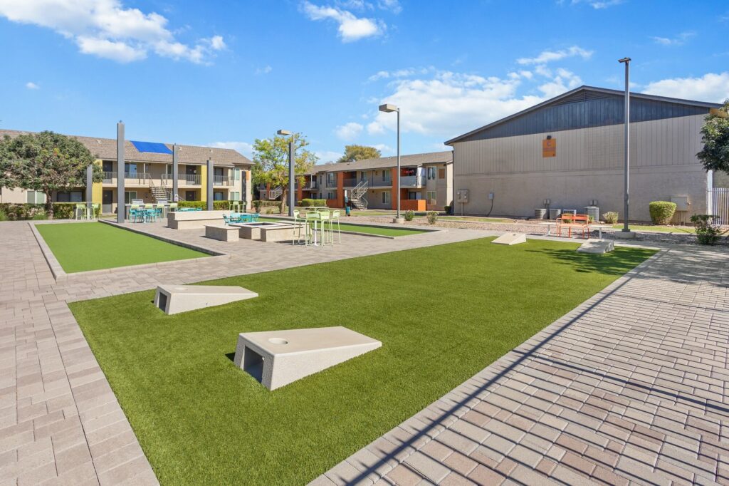 Outdoor recreation area with artificial turf, cornhole boards, seating, and tables between two apartment buildings under a bright blue sky with scattered clouds.