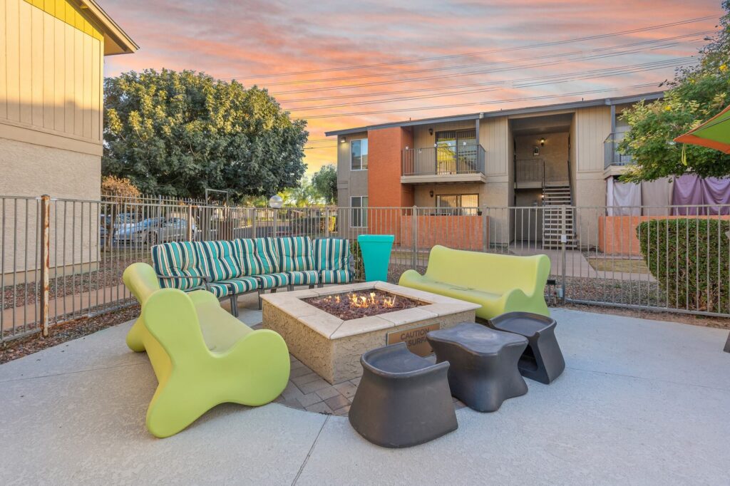 Outdoor patio with modern green and striped seating around a square fire pit, surrounded by metal fencing. Two apartment buildings and trees are visible in the background under a colorful sunset sky.