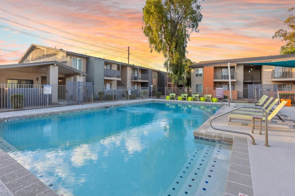Outdoor swimming pool surrounded by lounge chairs, with apartment buildings and a tree in the background. The sky is colorful with hues of orange and pink, suggesting sunset.