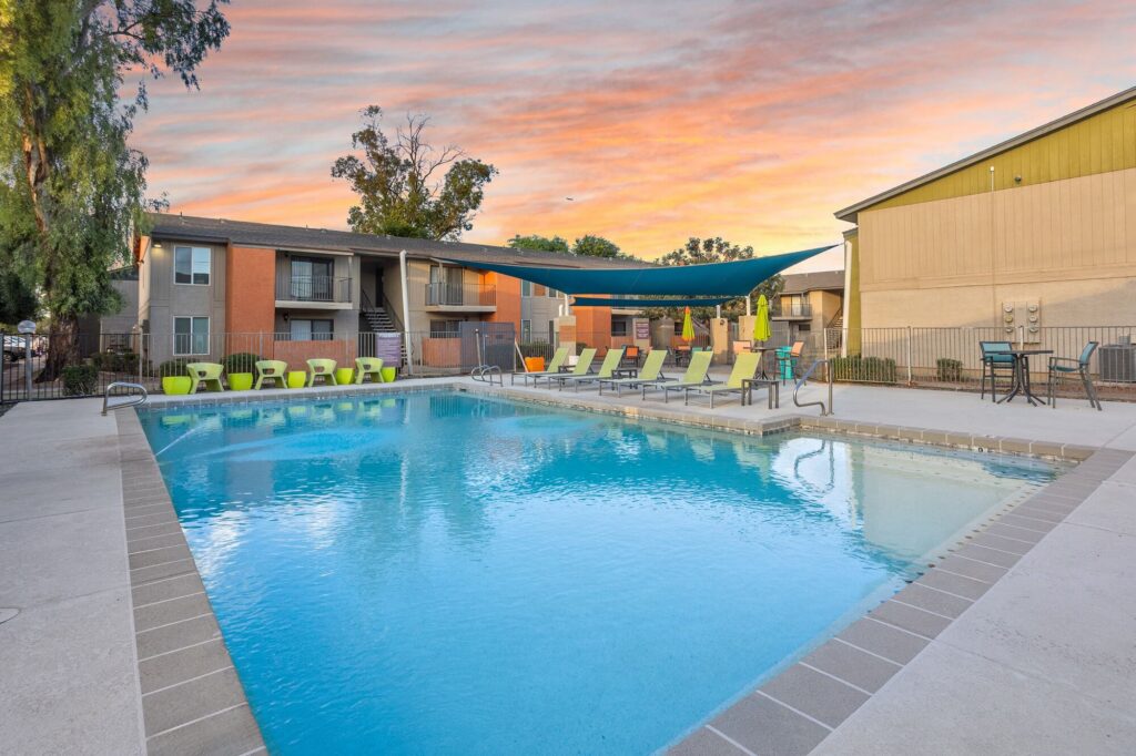 Apartment complex pool area at sunset, with lounge chairs, shade canopy, and surrounding buildings reflecting in the clear blue water.