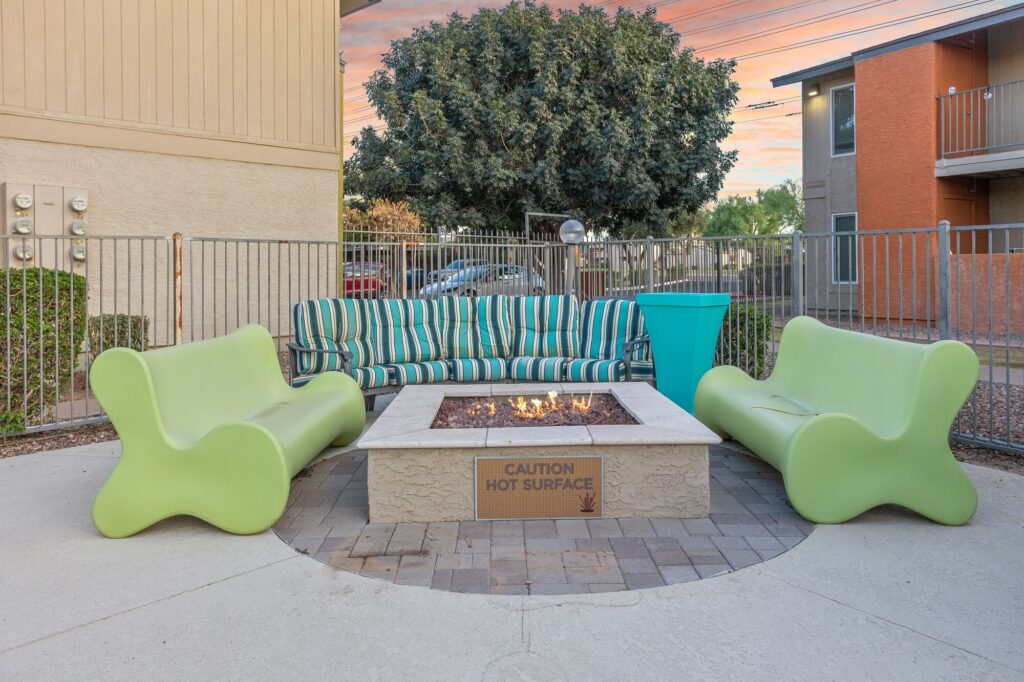 Outdoor seating area with two light green chairs, a striped sofa, and a fire pit in the center. A sign on the fire pit reads Caution Hot Surface. Theres fencing, trees, and buildings in the background.
