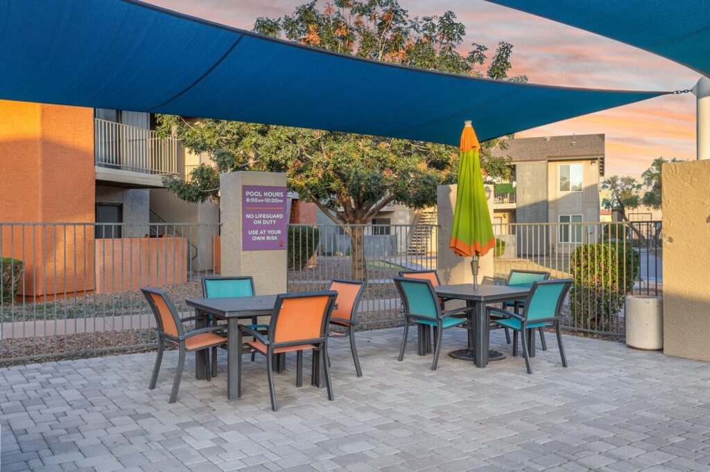 Outdoor patio with two tables and colorful chairs under blue shade sails. An umbrella stands between the tables. Apartment buildings and a tree are visible in the background. A sign with pool rules is posted near the fence.