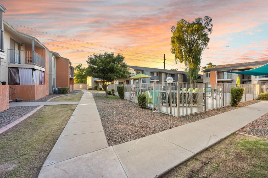 Apartment complex courtyard at sunset, featuring a fenced pool area with green lounge chairs, sidewalks, and surrounding buildings. Trees and a colorful sky are visible in the background.