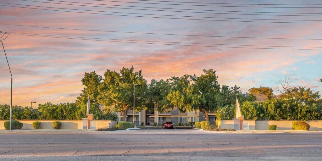 A modern building with trees and hedges in front, a red car parked in the center driveway, and a colorful sky with pink and orange clouds overhead. Power lines cross the upper part of the image.