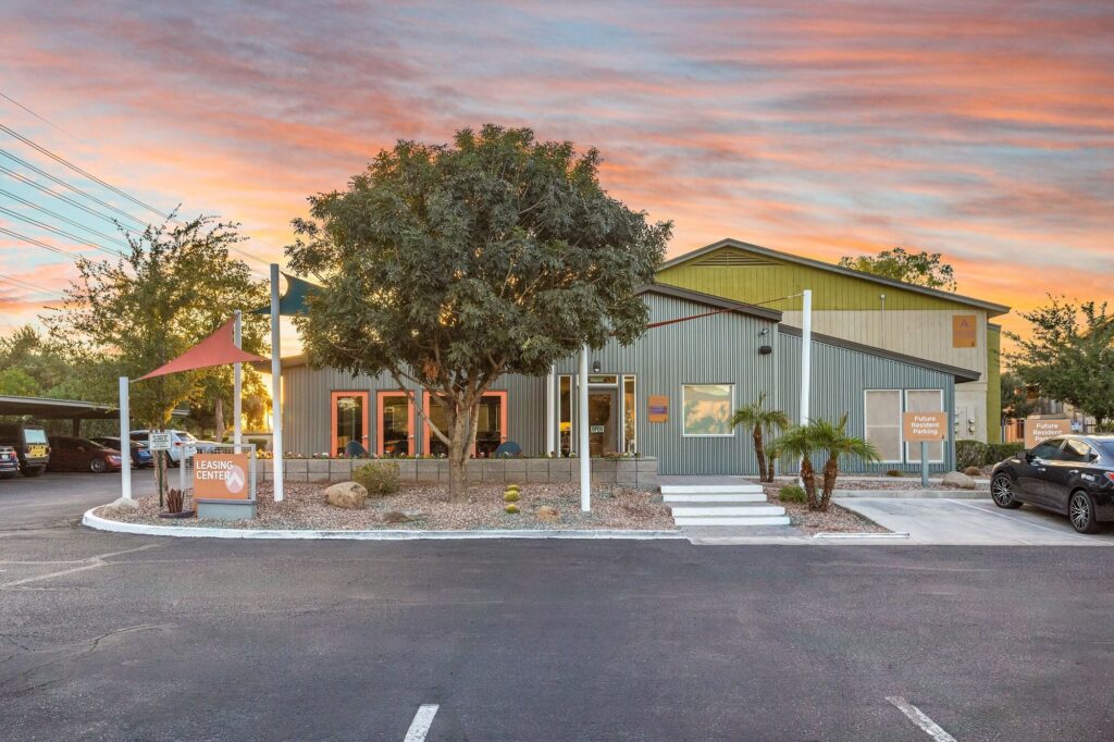 A modern building with large windows, outdoor seating, and a sign reading Learning Center. There are trees, a parking lot, and a colorful sunset sky in the background.