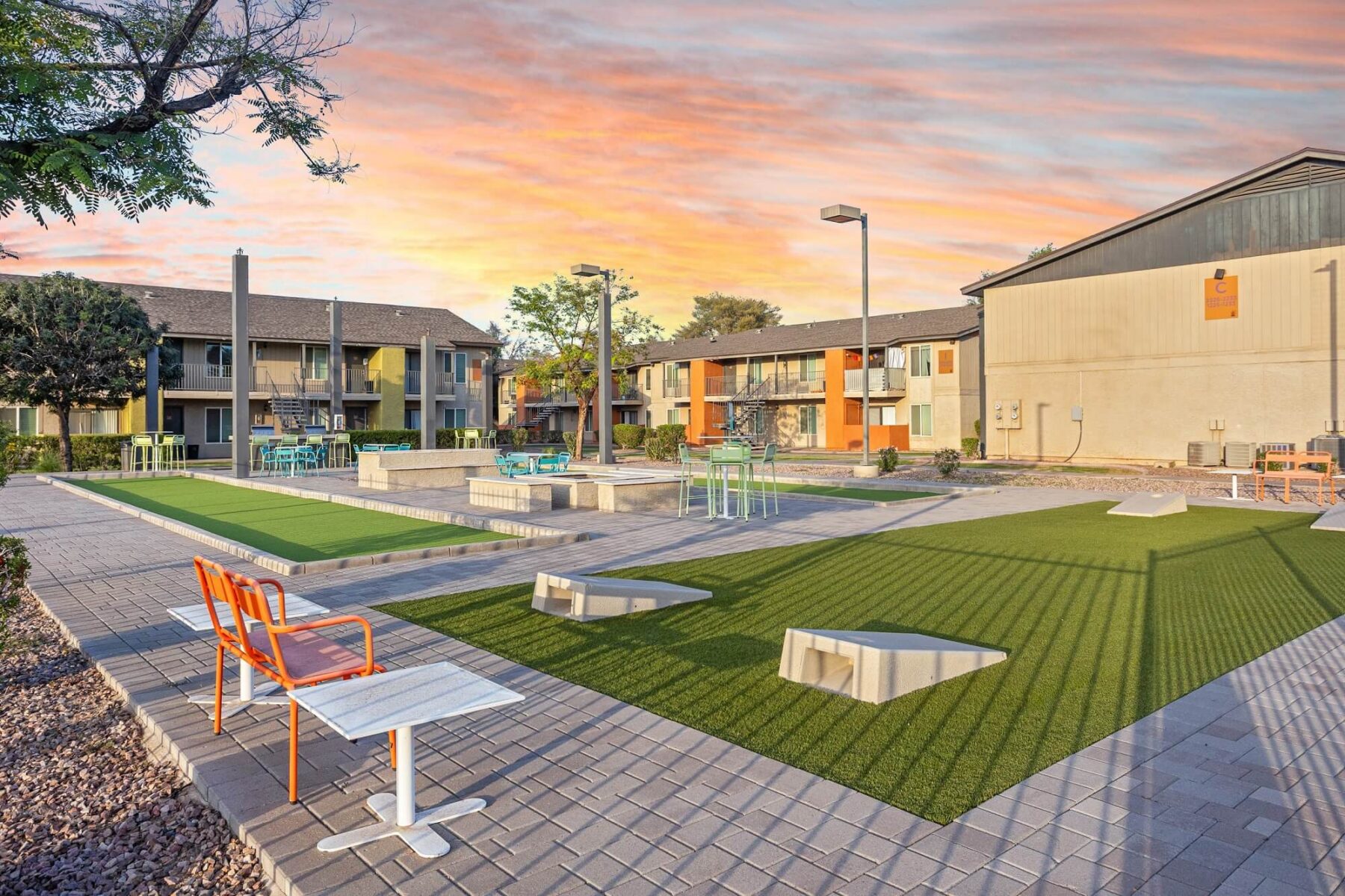Outdoor courtyard at an apartment complex features artificial turf, lounge chairs, tables, and walking paths, with modern buildings and a colorful sunset sky in the background.