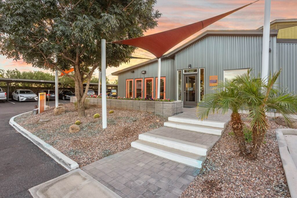Modern building exterior with corrugated metal walls, orange accents around windows, a sloped walkway, desert landscaping with rocks and cacti, and a red sunshade above the entrance. Cars are parked under a carport nearby.