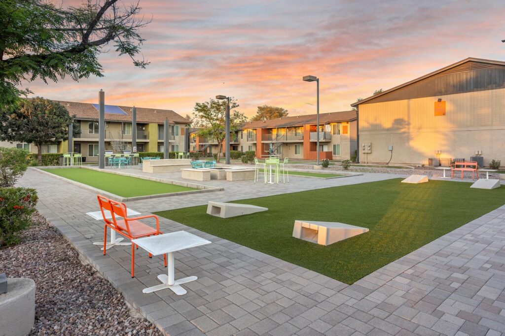 Modern apartment courtyard at sunset with artificial turf, orange and white benches, outdoor seating, and cornhole boards on a paved area, surrounded by buildings and landscape.