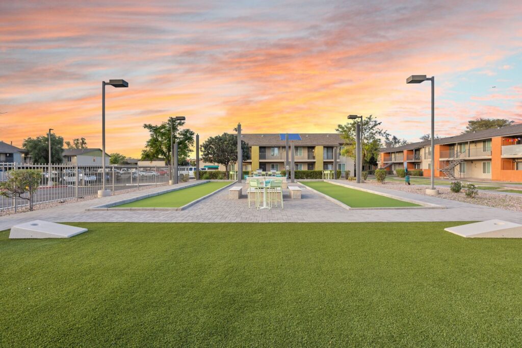 Outdoor recreation area with two bocce ball courts, a table with chairs, neatly trimmed artificial grass, and apartment buildings in the background under a colorful sunset sky.