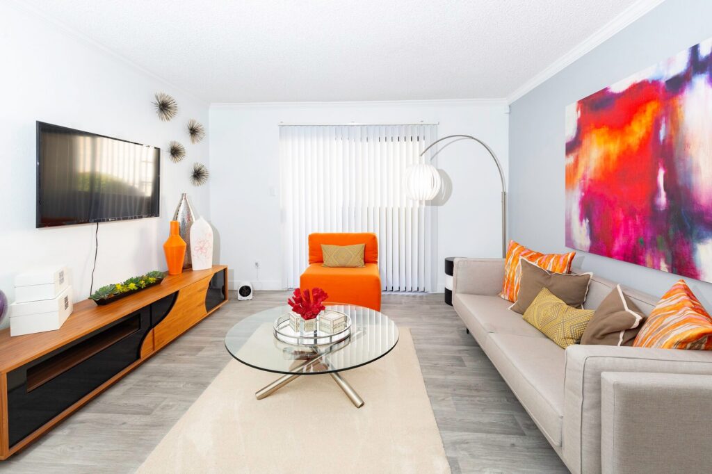 Modern living room with a gray sofa, orange chair, glass coffee table, TV on the wall, stylish decor, and colorful abstract art above the sofa. Vertical blinds cover the window, and a curved floor lamp stands nearby.