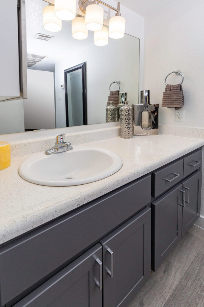 Modern bathroom vanity with gray cabinets, a white countertop, a round sink, silver faucet, mirror, towel ring with brown towel, decorative vases, and wall-mounted lights above the mirror.