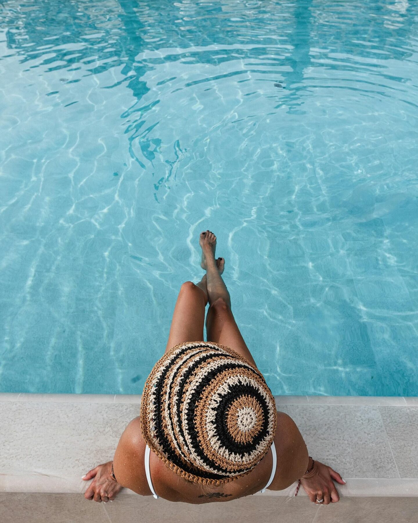 A person wearing a crocheted hat and white swimsuit sits at the edge of a clear blue pool with their legs in the water, enjoying a relaxing day at one of the pet friendly apartments, viewed from above.