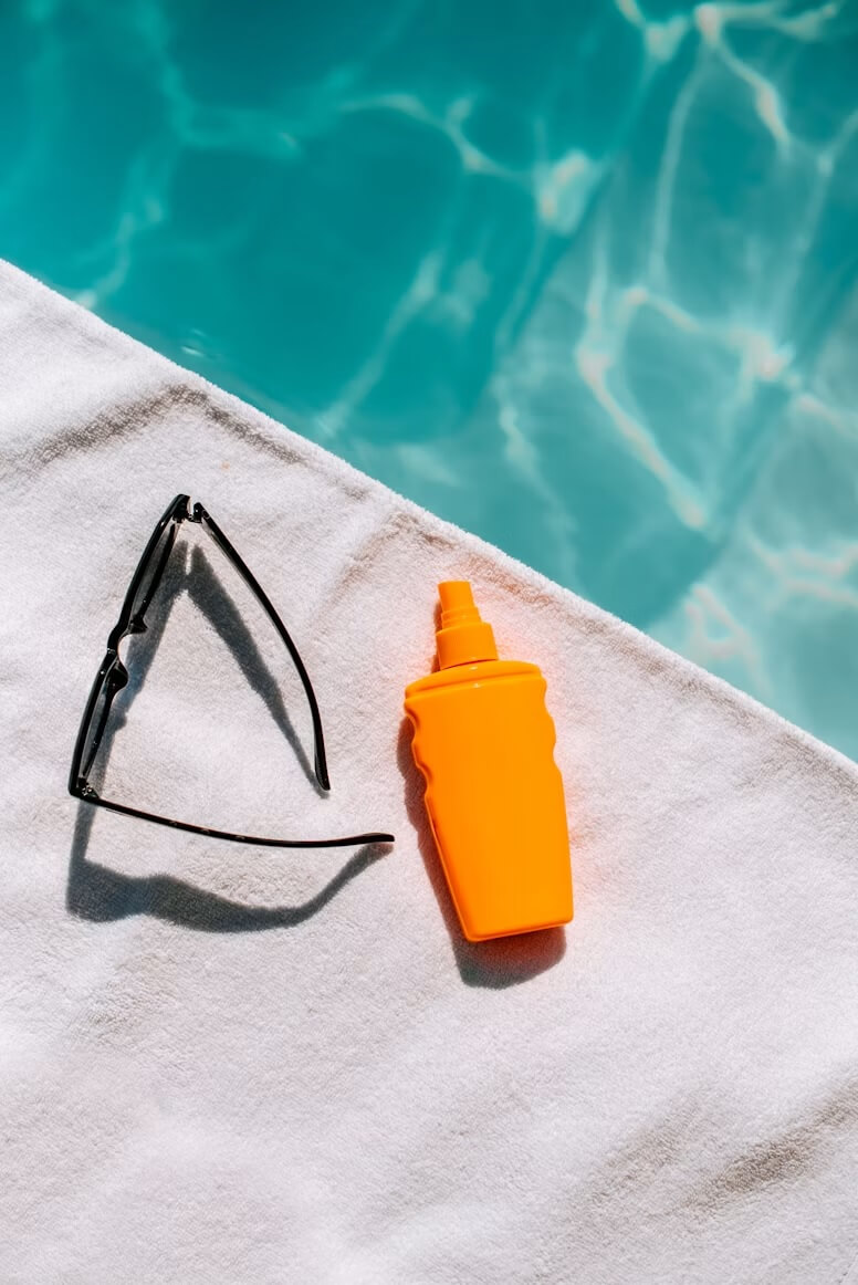 Sunglasses and an orange sunscreen bottle on a white towel beside a clear blue swimming pool.
